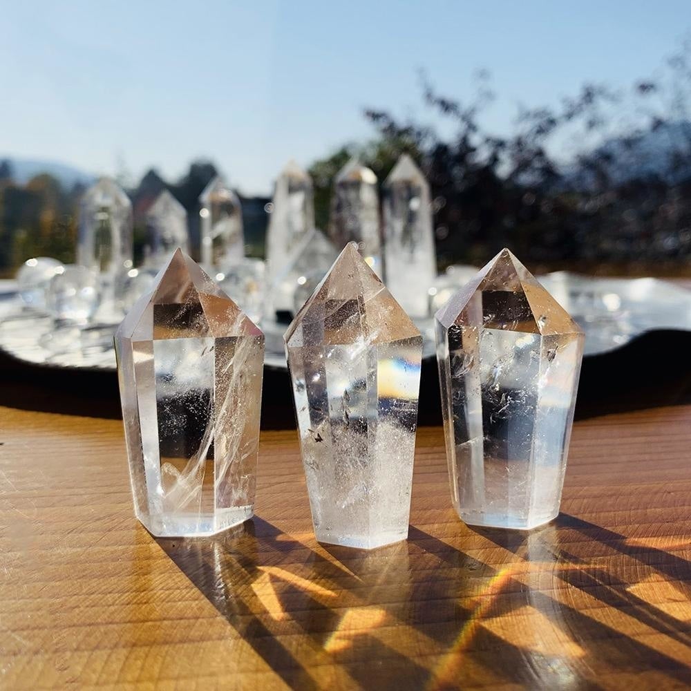 Three clear quartz wands with six-faceted tips standing in sunlight on a wooden table, casting light and a small rainbow