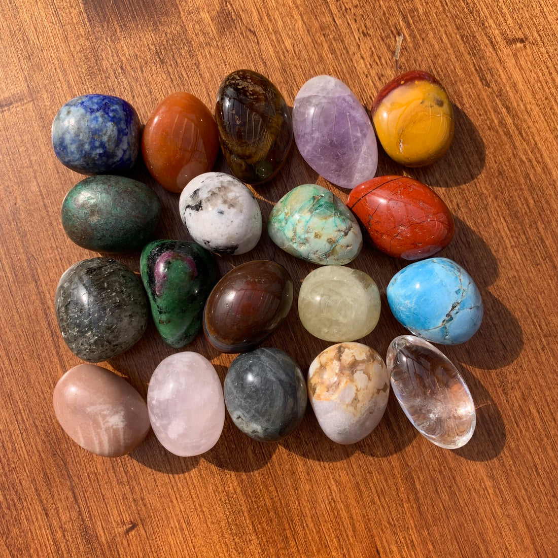 All nineteen tumbled stones laid out in rows on a wooden table: lapis, amethyst, mookaite, red jasper, apatite, clear quartz, rose quartz and more