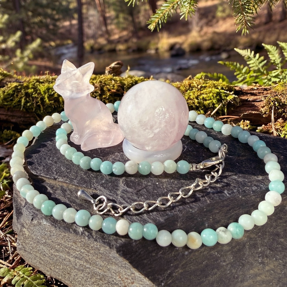 The rose quartz fox beside the rose quartz sphere resting on the pink agate ring, circled by the Caribbean jasper necklace on a slate rock in the forest