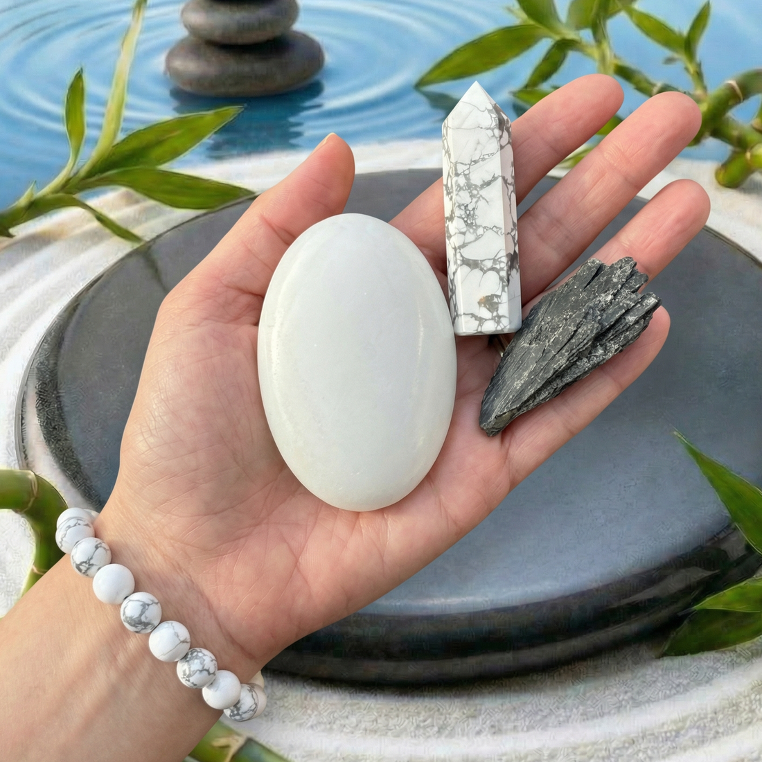 An open hand holding a white oval palmstone, a white-and-grey veined howlite point and a black kyanite fan, with a howlite bead bracelet on the wrist