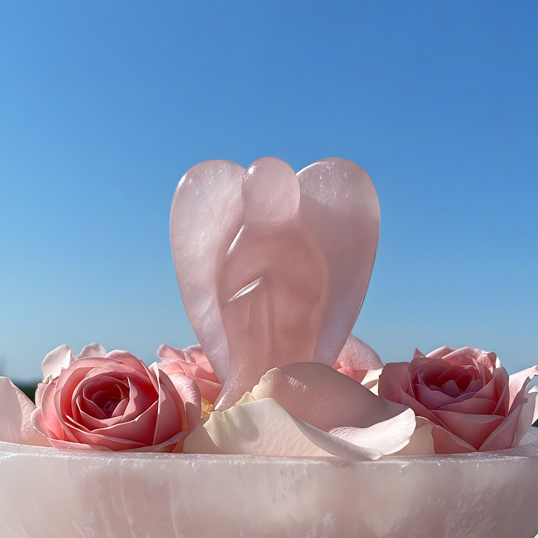 Rose quartz angel carving with folded wings, set among pink roses under a blue sky