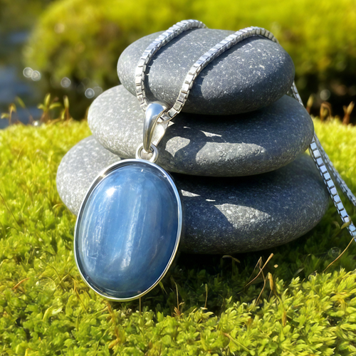 An oval blue kyanite pendant with blade-like streaks in a sterling silver bezel on a silver box chain, draped over stacked stones on moss