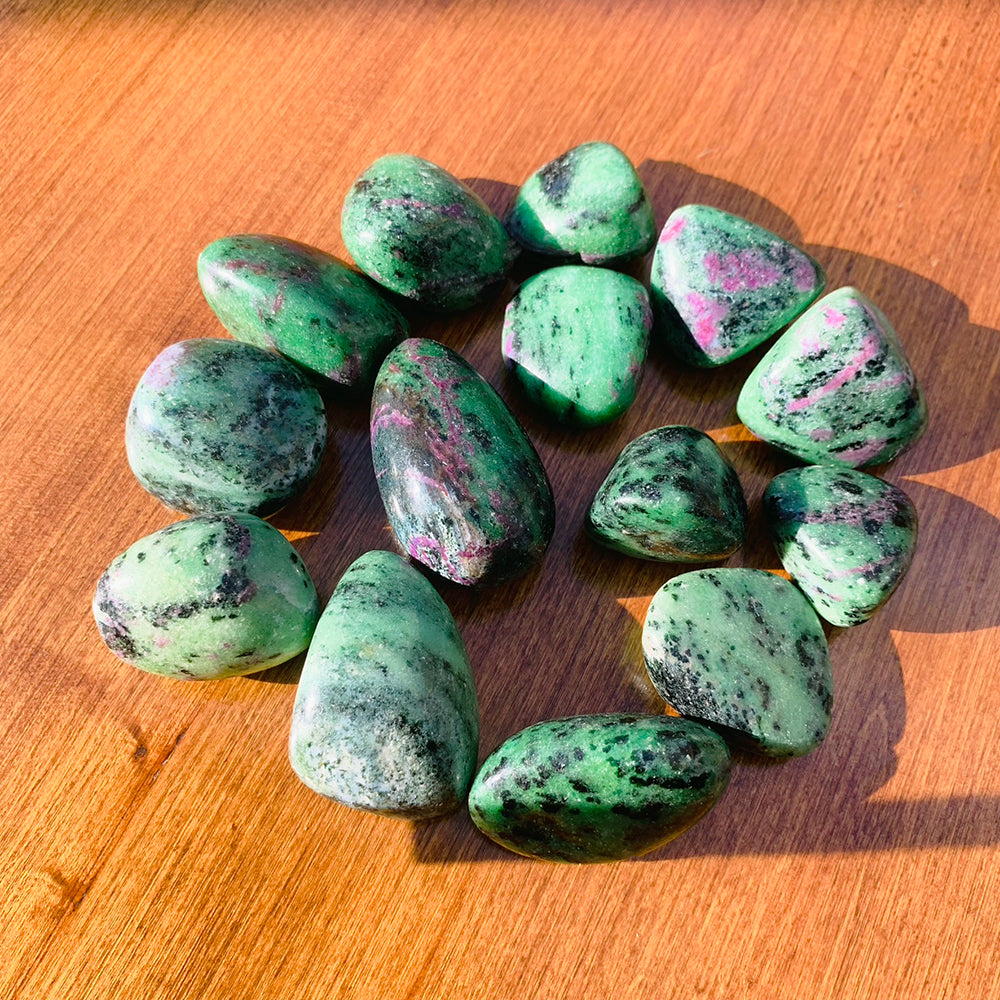 A ring of polished green ruby in zoisite tumbled stones with black flecks and magenta ruby patches on a sunlit wooden table; each order includes one stone