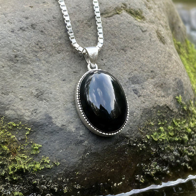 A polished black obsidian oval pendant in a beaded sterling silver bezel on a silver box chain, resting on a mossy stone