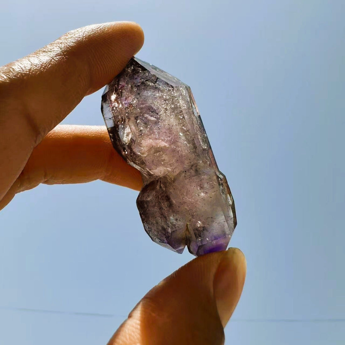 The Super 7 freeform held up against a blue sky, smoky and lilac quartz with fine dark inclusions and a deep purple tip