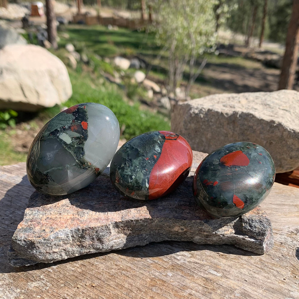 Three African bloodstone palmstones on a slab of rock outdoors, each with its own mix of green, grey and red
