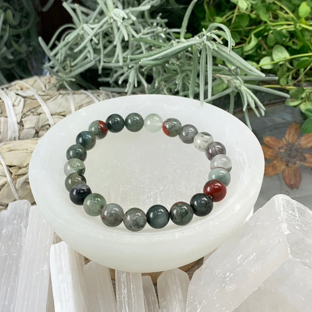 An African bloodstone bead bracelet in deep green and grey with red patches, resting in a selenite bowl