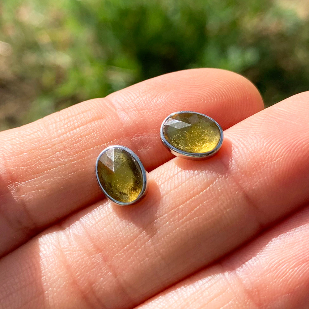 Two oval faceted honey-gold amber studs in sterling silver bezels resting on fingertips in sunlight
