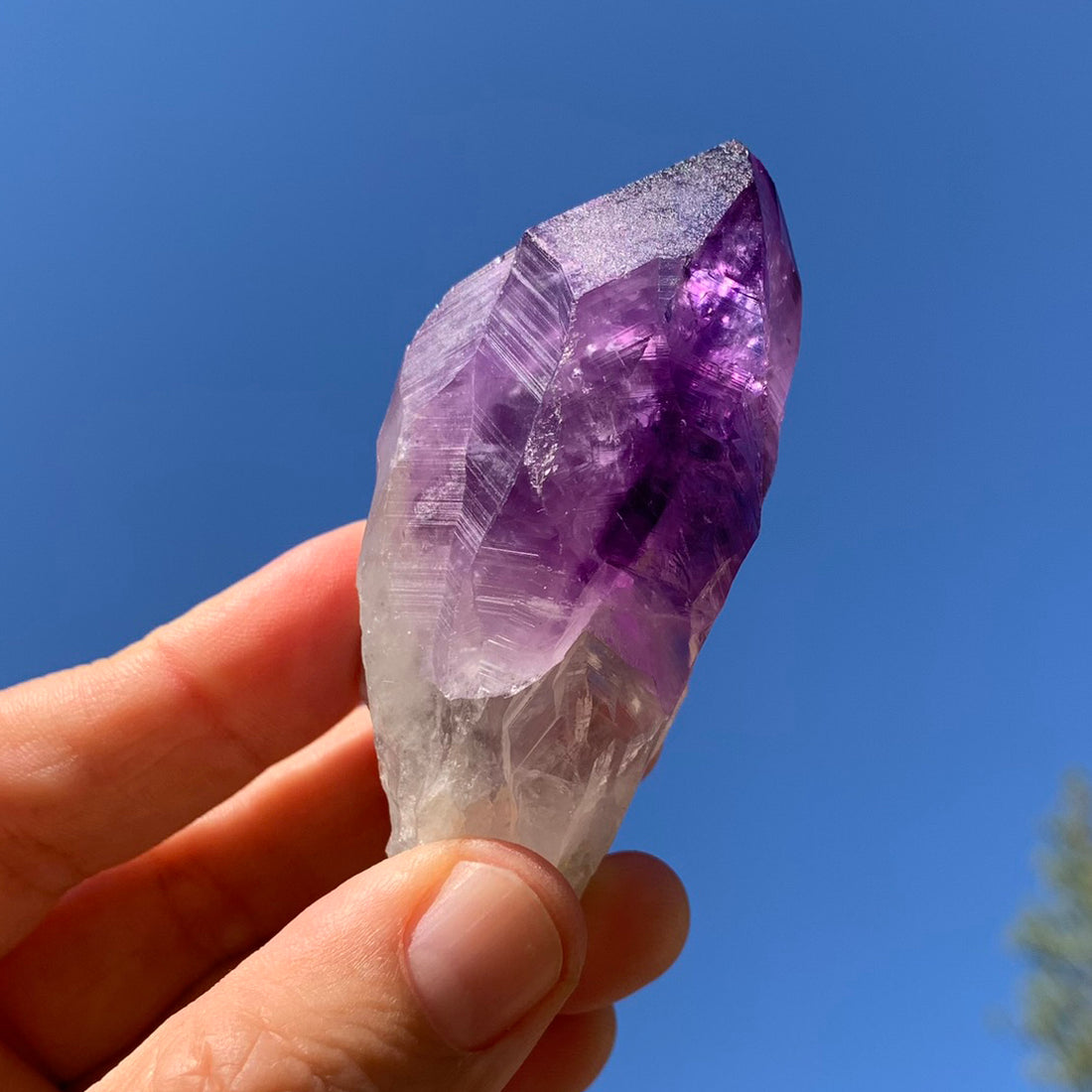 A raw amethyst shard held up to a blue sky, deep violet at the tip and clear at the base