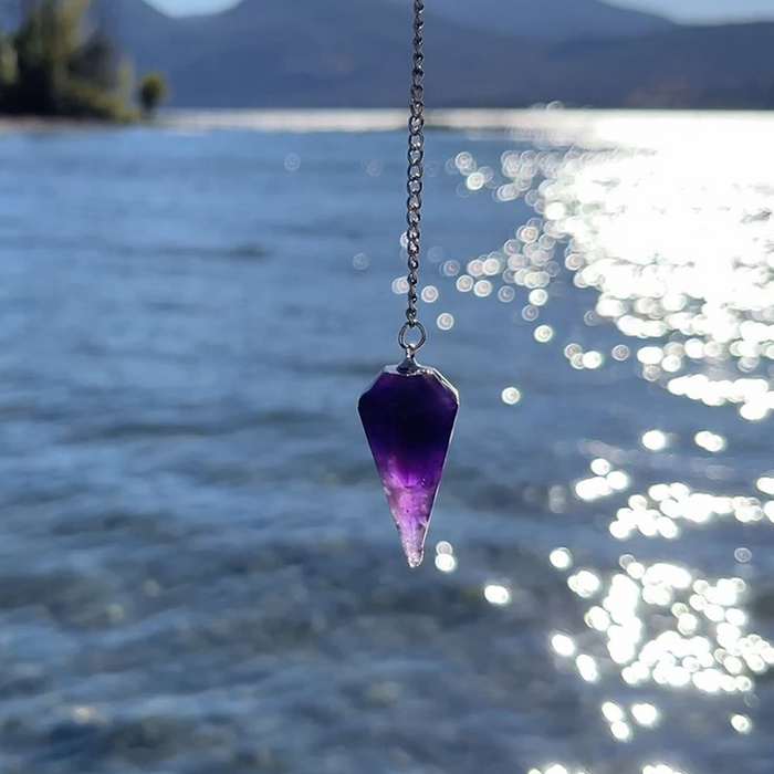 A faceted amethyst pendulum hanging on a silver chain above sparkling water