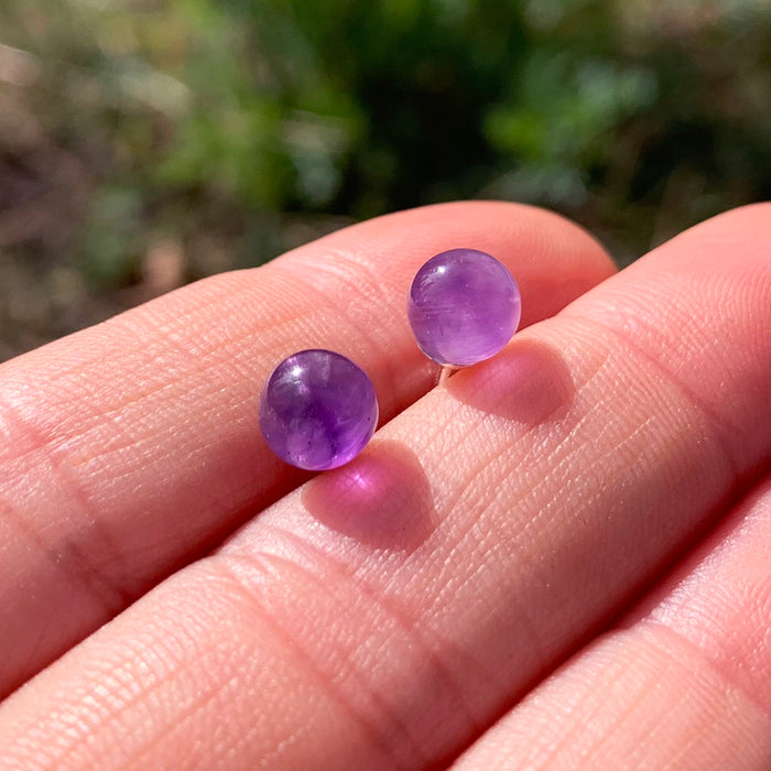 A pair of round polished amethyst stud earrings resting on fingertips in sunlight