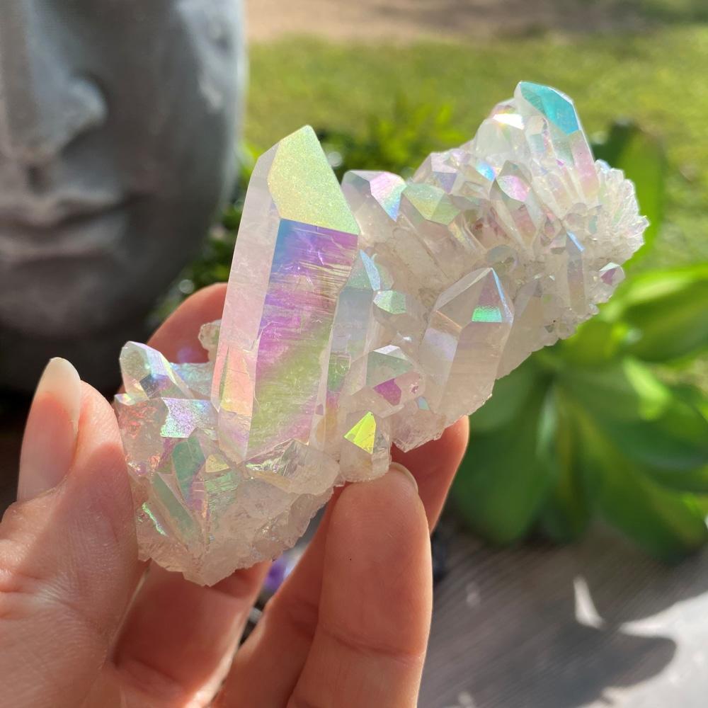 An angel aura quartz cluster held in the fingers, its points shimmering pink, gold, green and blue in the sun