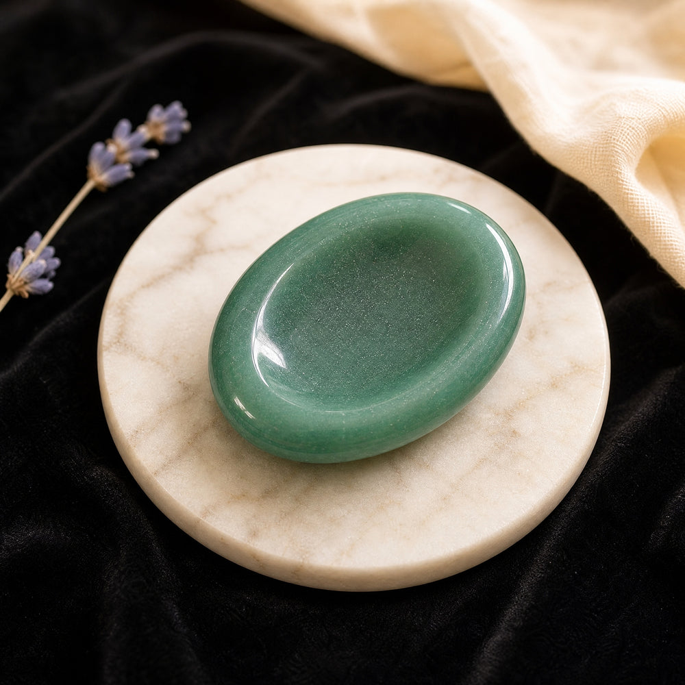 A polished green aventurine worry stone with a smooth thumb hollow, resting on a round marble dish beside lavender