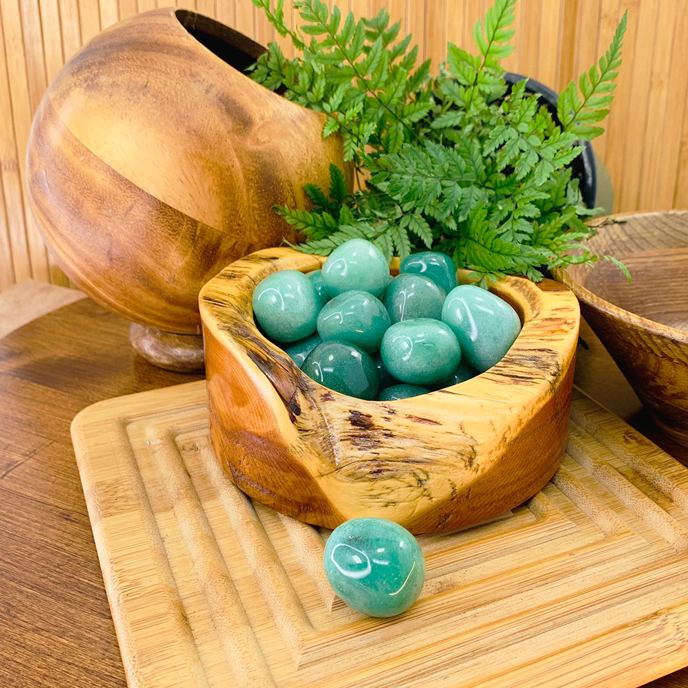 A wooden bowl of polished green aventurine tumbled stones beside ferns, with one stone resting in front