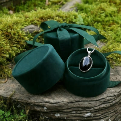 The black obsidian pendant tucked into an open round green velvet jewelry box on a mossy stone