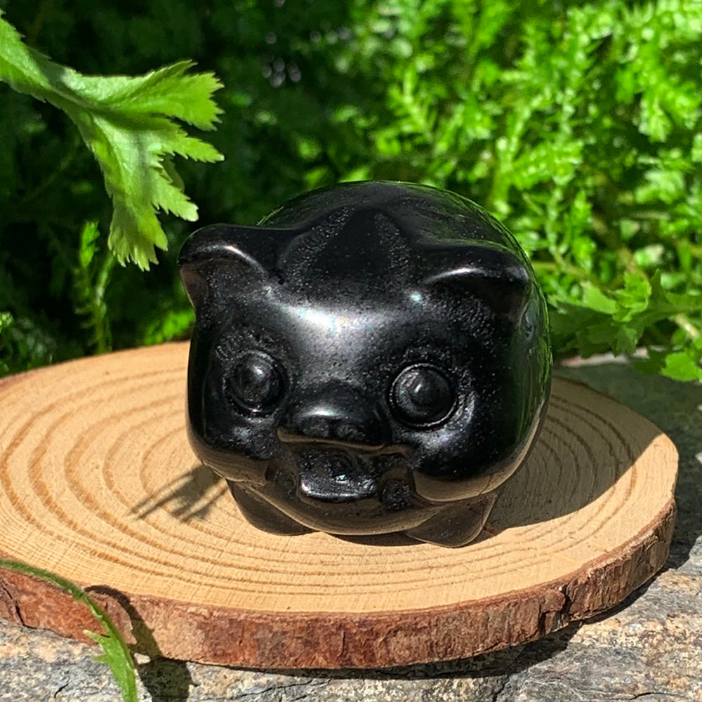 A round, glossy black agate pig totem with little ears and snout, sitting on a wooden slice in front of ferns