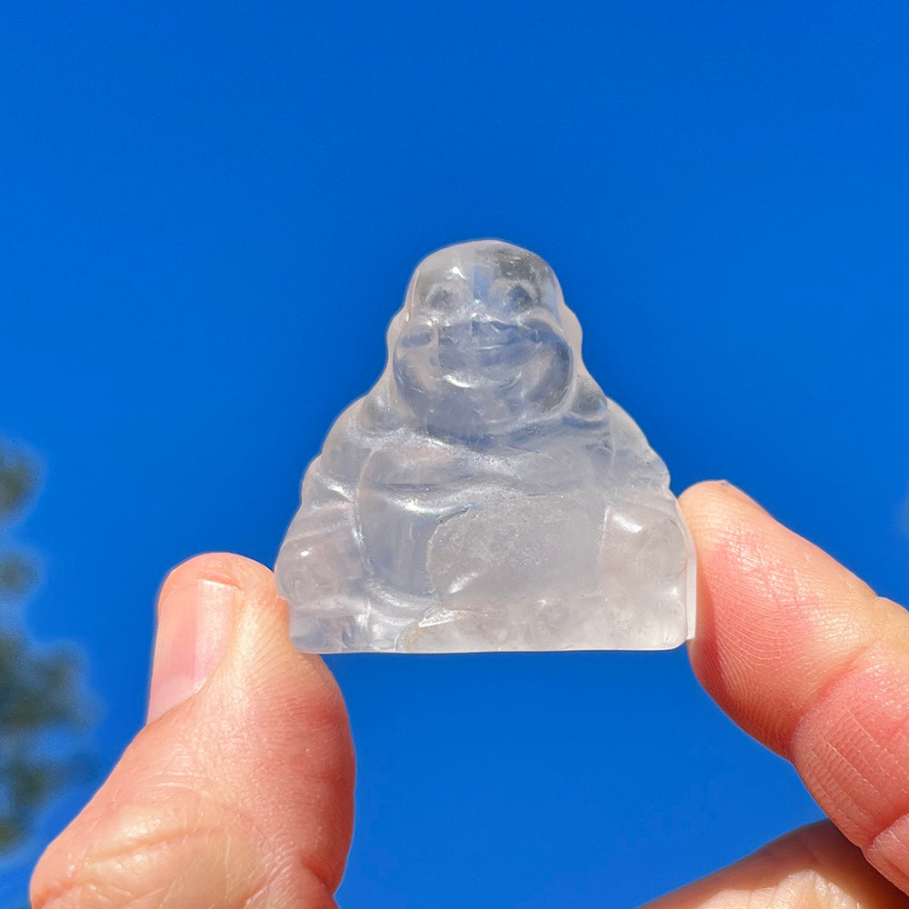 The small clear quartz Buddha held between fingertips against a bright blue sky
