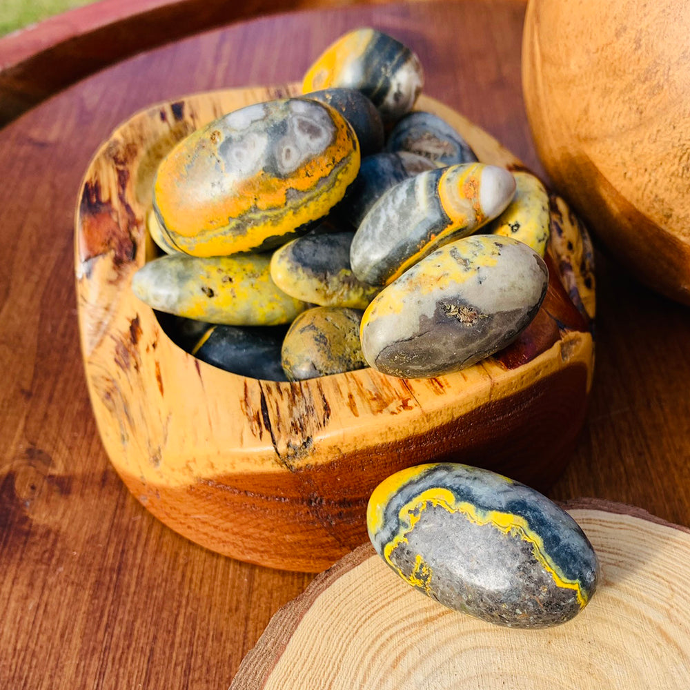 Polished bumblebee jasper tumbled stones banded in bright yellow, orange, grey and black, piled in a wooden bowl with one stone resting on a wood slice