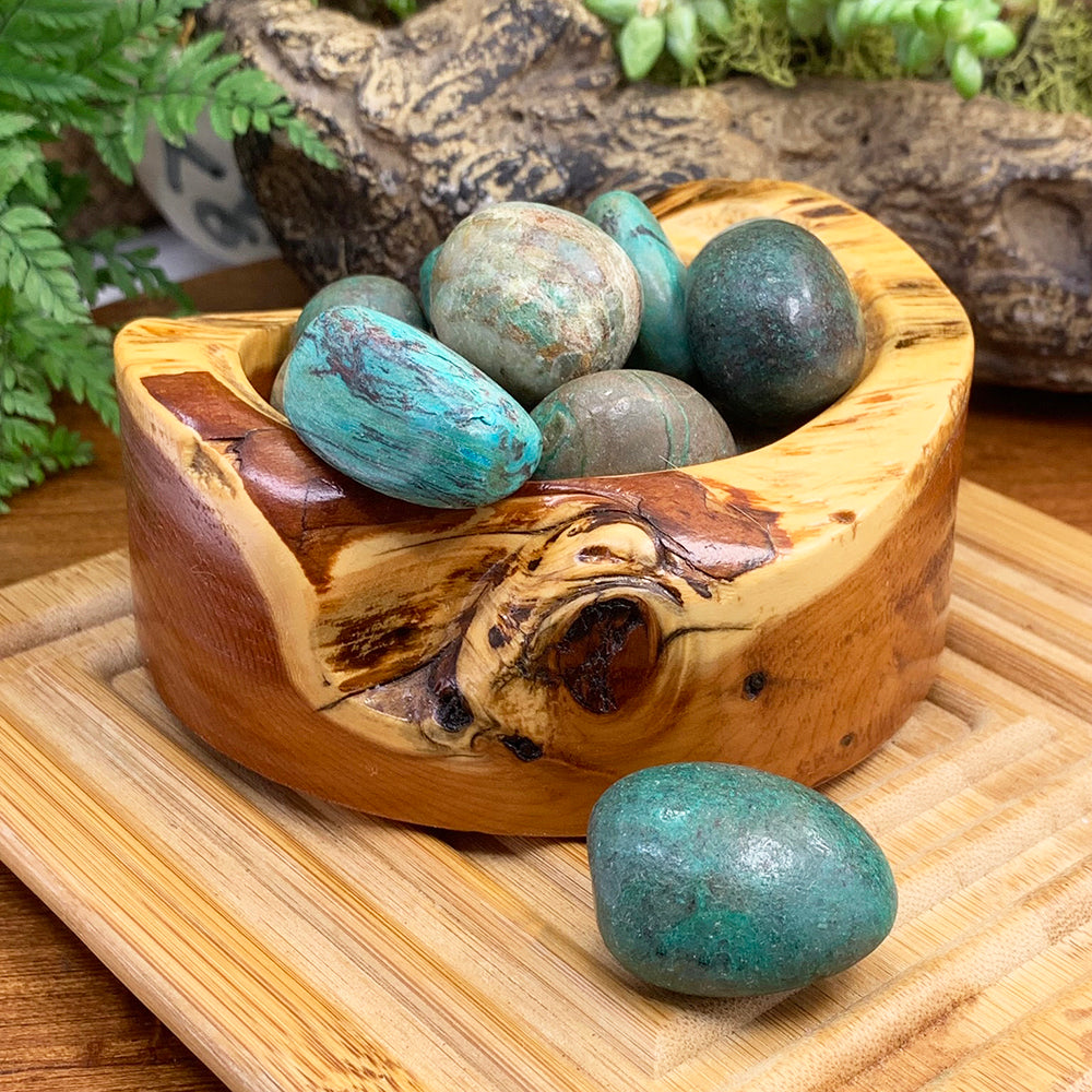 Polished chrysocolla tumbled stones in turquoise, teal and sea green in a carved wooden bowl, with one resting in front on a bamboo tray