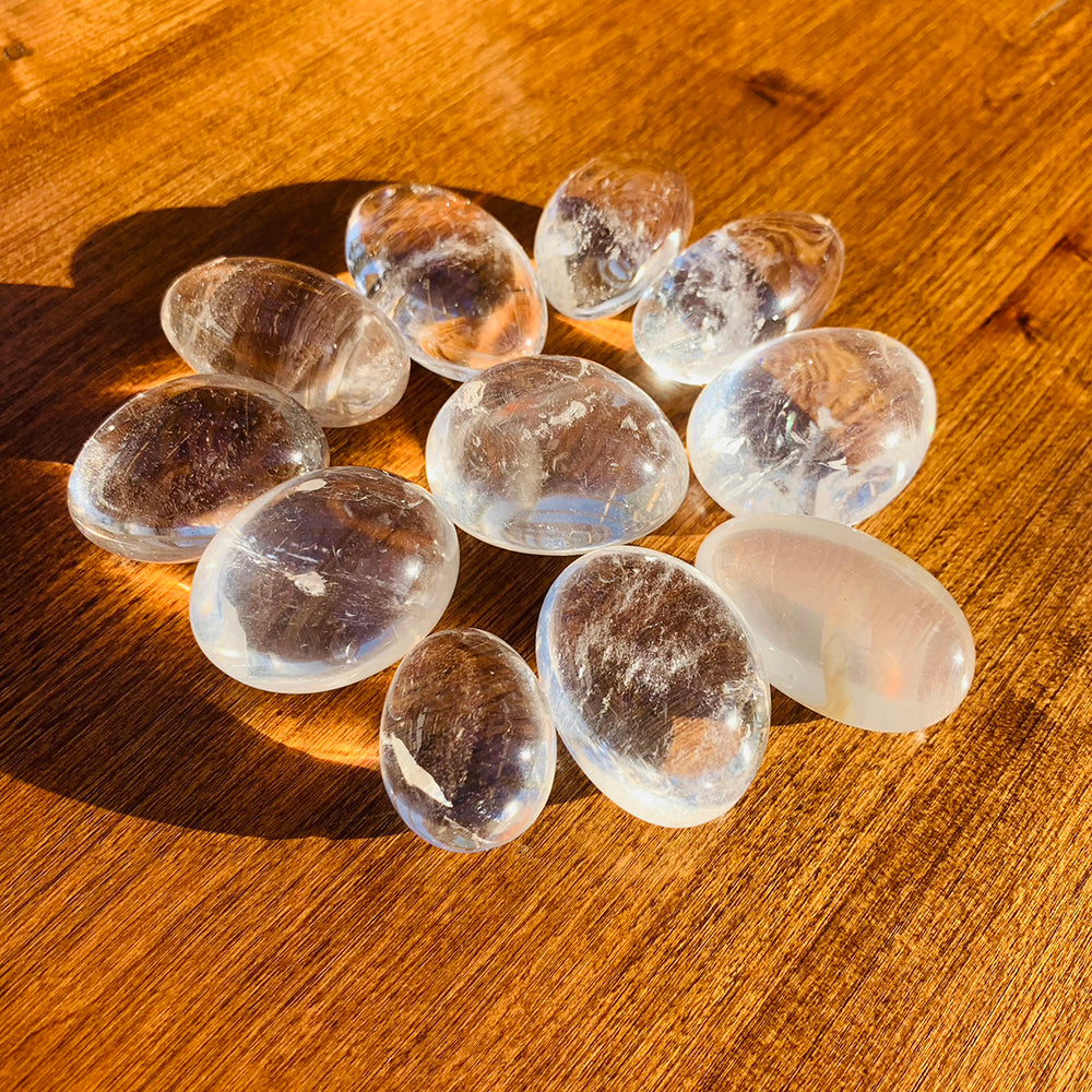 A dozen clear quartz ellipse stones in warm sunlight on a wooden table, each with its own inner veils and clarity