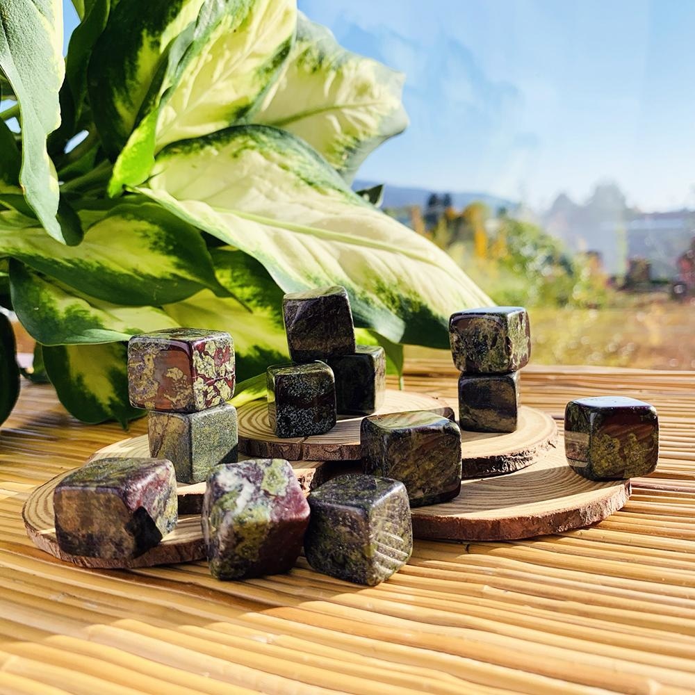 Polished dragon&rsquo;s blood jasper tumbled cubes in deep green swirled with blood red, stacked on wood slices in sunlight beside a leafy plant
