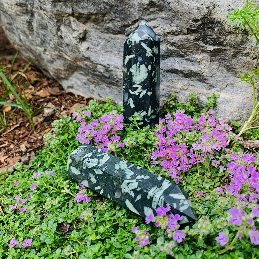 Two polished flower quartz agate points, dark stone scattered with pale flower-like patterns, one standing and one lying among purple thyme flowers