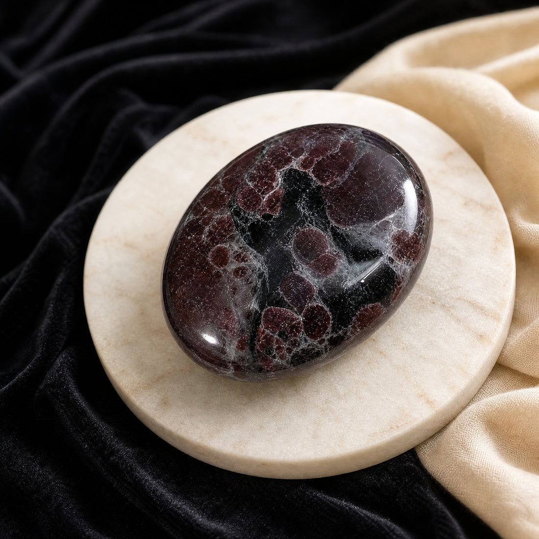 Polished red garnet palmstone with wine-red garnet patches and black veining on a pale marble dish