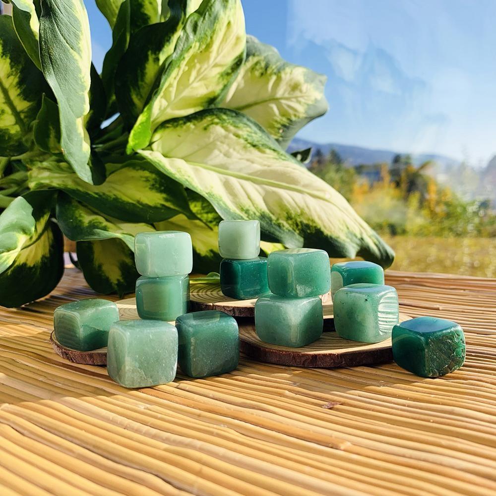 Green aventurine tumbled cubes in light and deep greens, stacked on wood slices in sunlight beside a leafy plant