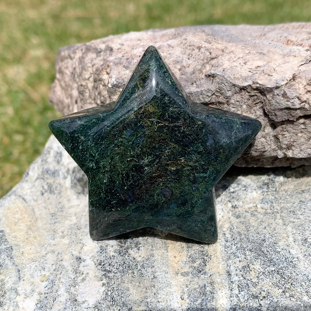 A polished five-pointed moss agate star in deep green, patterned with fine moss-like threads, resting against a sunlit rock