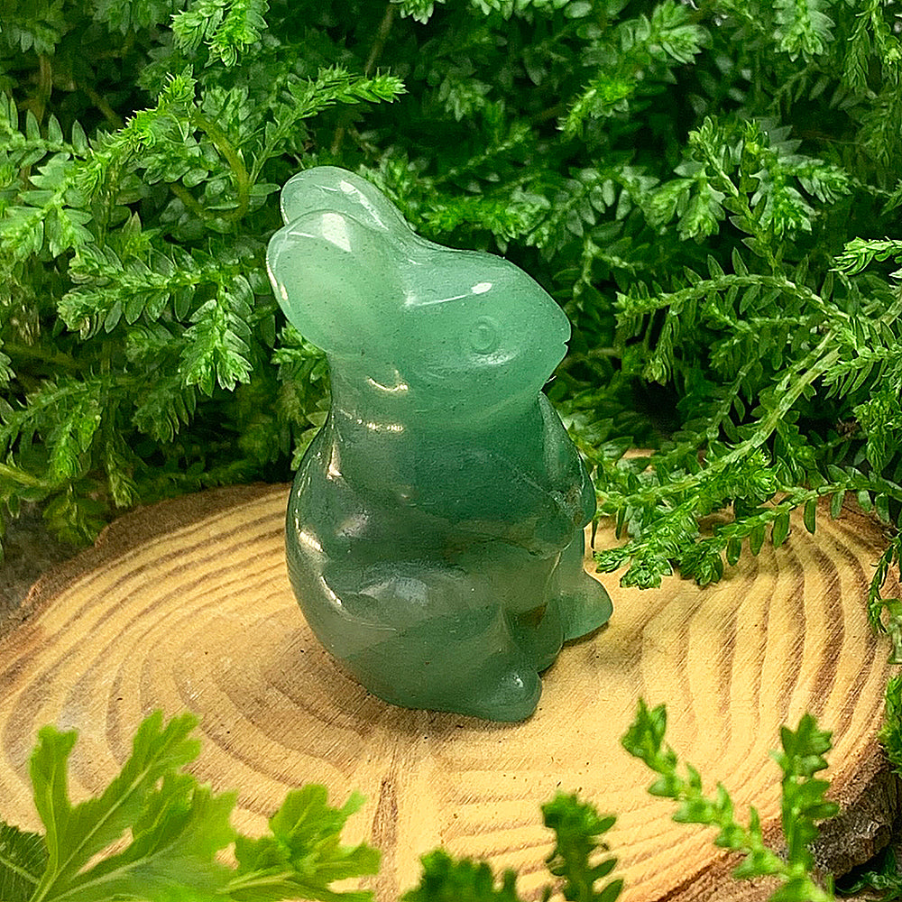 A small carved green quartz rabbit sitting upright on a wooden slice among ferns