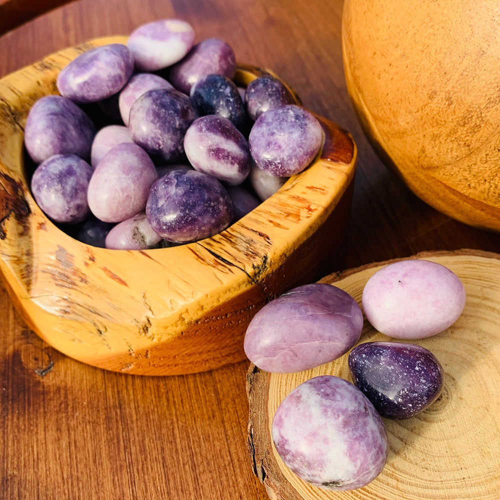 A wooden bowl of lepidolite tumbled stones in shades of lilac, lavender and deep purple, with a few on a wood slice