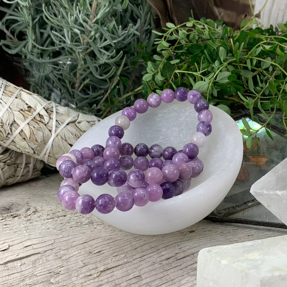 Lilac and violet lepidolite bead bracelets resting in a white selenite bowl beside sage bundles and herbs