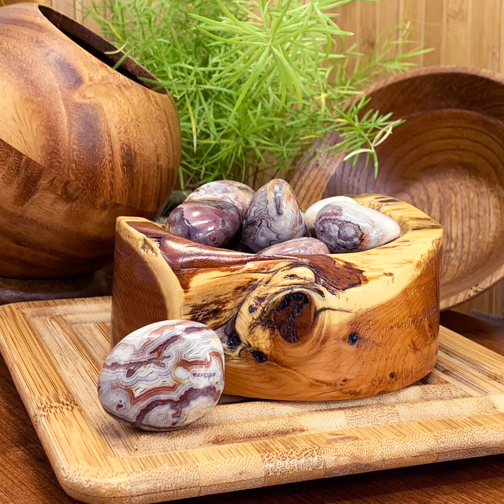A polished Mexican lace agate ellipse with swirling red, grey and cream lace bands resting in front of a carved wooden bowl of more lace agate stones