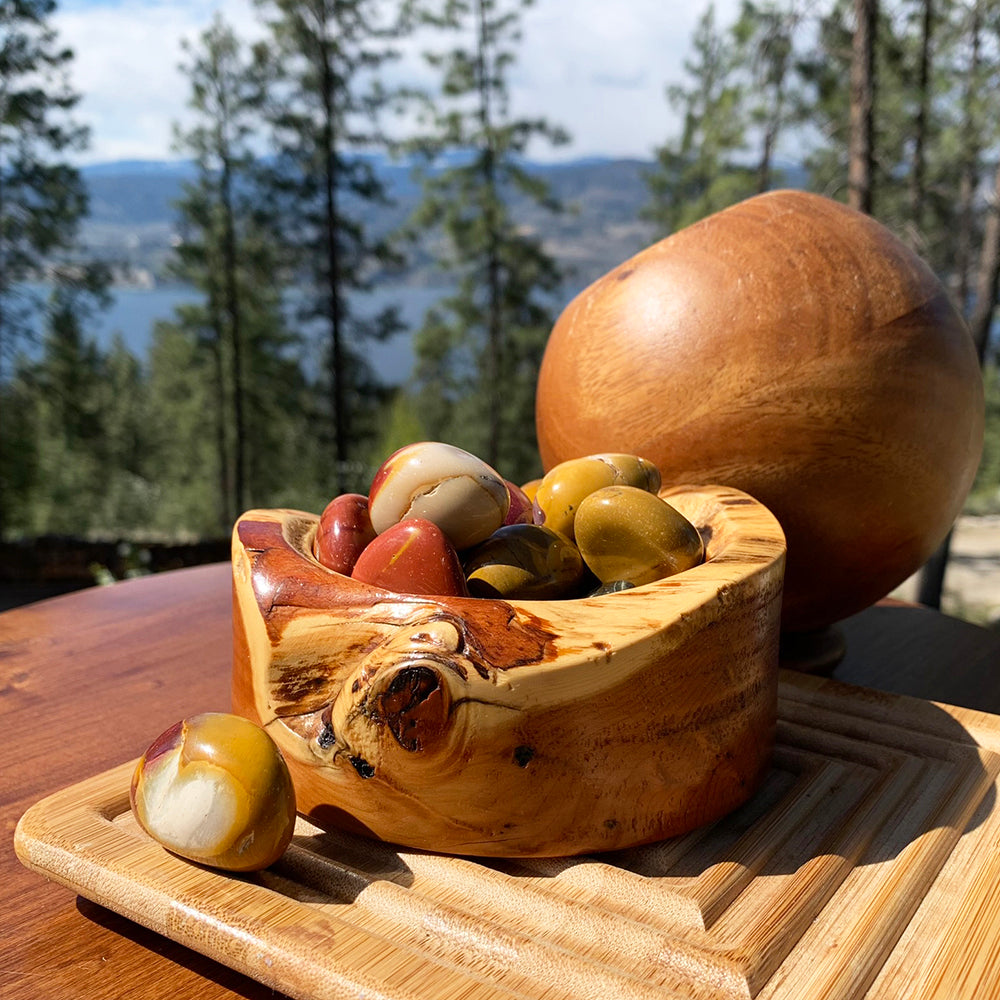 Polished mookaite tumbled stones in mustard yellow, deep red and purple, piled in a burl wood bowl with one stone resting in front