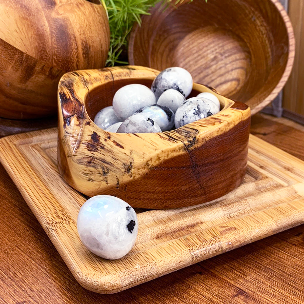 A wooden bowl of tumbled moonstone ellipses with one ellipse resting on a bamboo tray in front