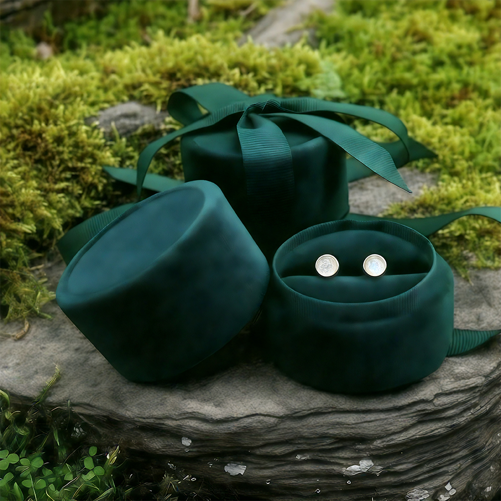 The pair of moonstone studs set into an open round green velvet jewelry box on a mossy stone, beside its lid and a ribboned velvet box