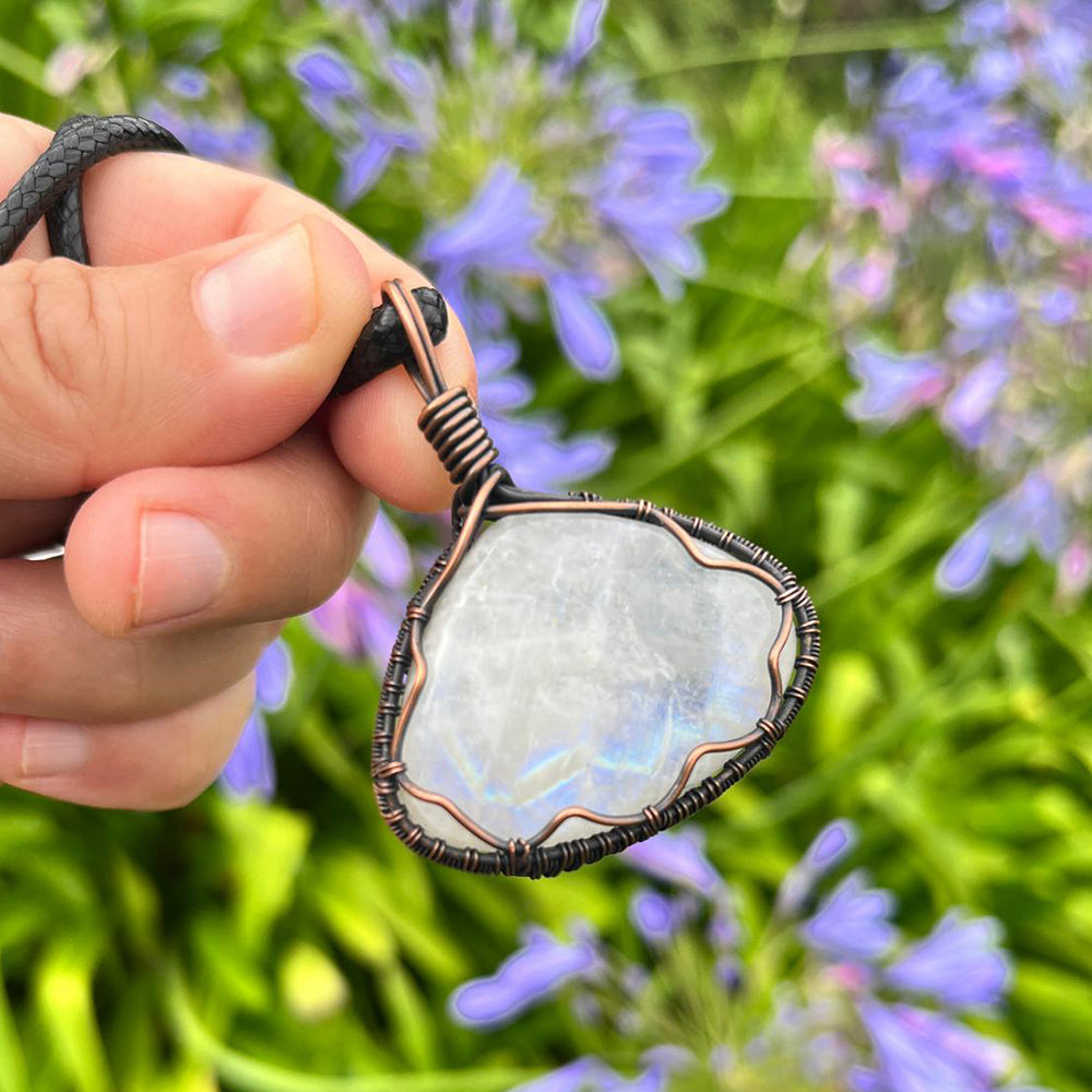 Fingers holding the moonstone pendant by its black braided cord in front of purple agapanthus flowers