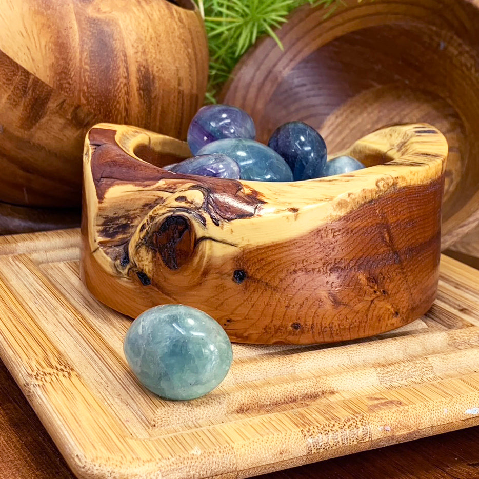 A tumbled sea-green fluorite ellipse resting in front of a wooden bowl of purple, blue and green fluorite stones