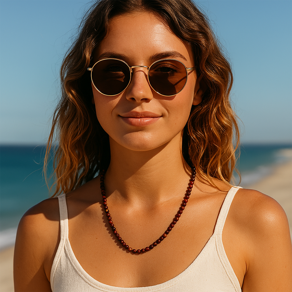 A woman in round sunglasses and a white tank top wearing the red obsidian bead necklace on the beach