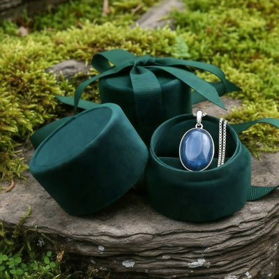 The blue kyanite pendant on its chain inside an open round green velvet jewelry box on a mossy stone