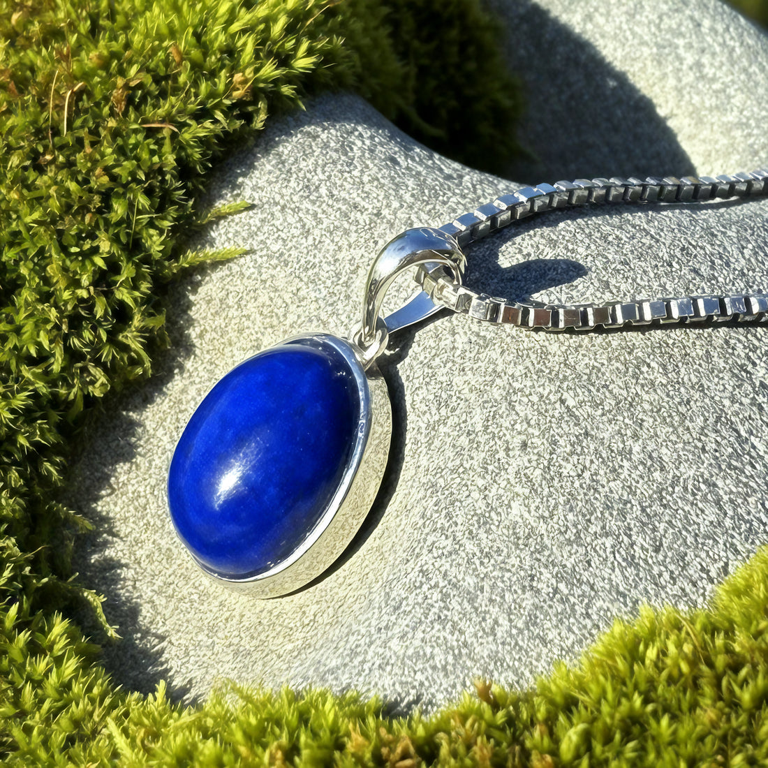 Close-up of the oval lapis lazuli pendant in a sterling silver bezel, resting on a mossy stone