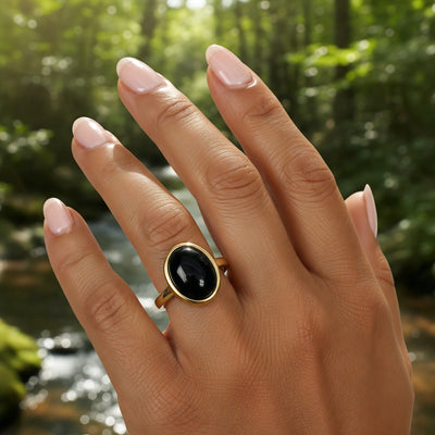 The black onyx and gold ring worn on a hand in front of a forest stream