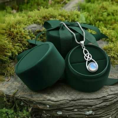 The moonstone infinity knot necklace resting in an open round green velvet jewelry box on a mossy stone