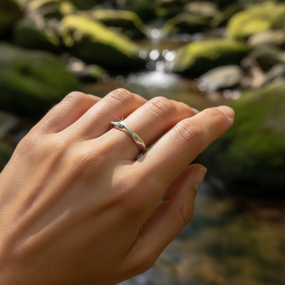 The silver wave ring worn on a finger beside a mossy stream, showing its slim, wavy profile on the hand