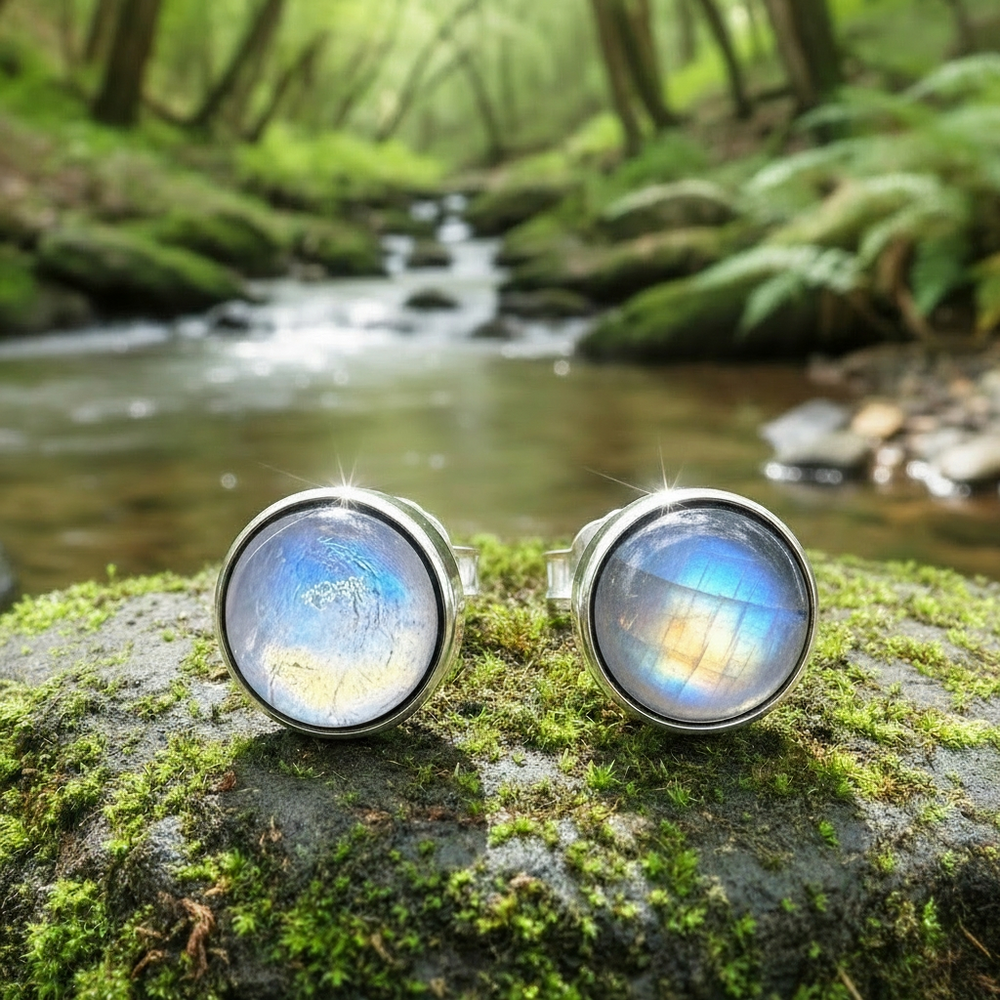 Close-up of the two round moonstone studs glowing blue-white in polished silver bezels, resting on a mossy stone beside a forest stream