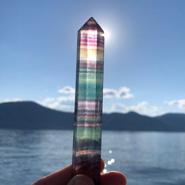 The rainbow fluorite point generator held up against a bright sky over the water, its violet and green bands glowing