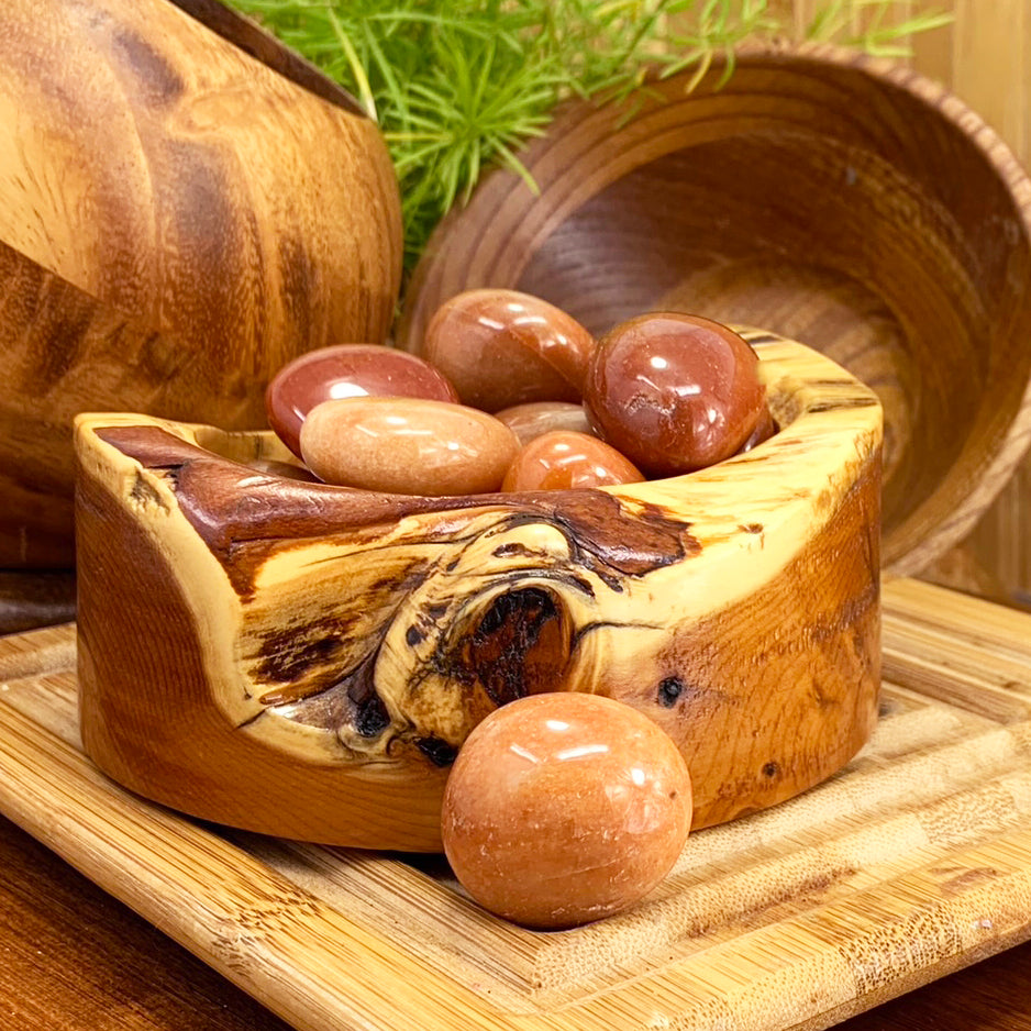 Polished red aventurine ellipses in shades of peach, rust and deep red, piled in a carved wooden bowl with one resting in front