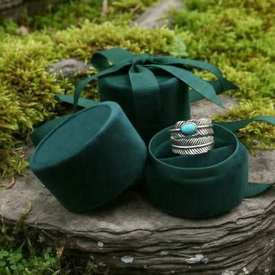 The feather ring nestled in an open round green velvet ring box beside its lid and ribbon on a mossy stone