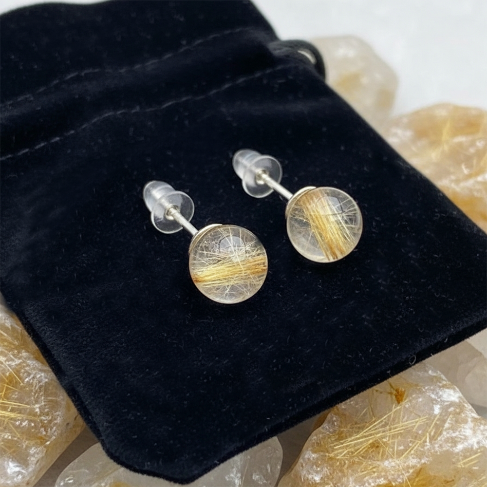 The pair of rutilated quartz studs with their posts and clear backs, laid on a black velvet drawstring pouch among rough rutilated quartz