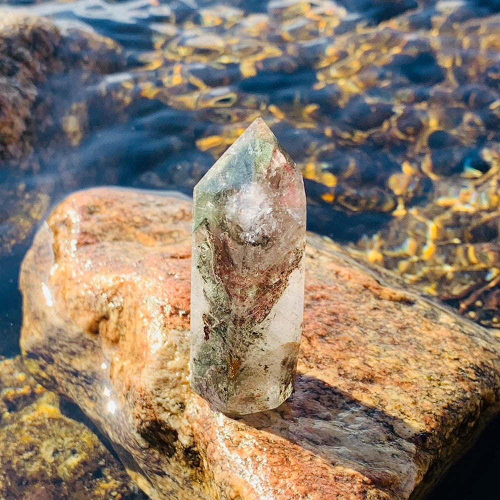 A clear Shaman Quartz point standing on a riverside rock, with green and red-brown mineral inclusions inside